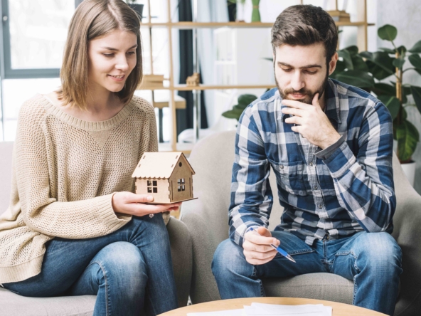 una mujer sostiene una casa de madera y un hombre calcula una hipoteca