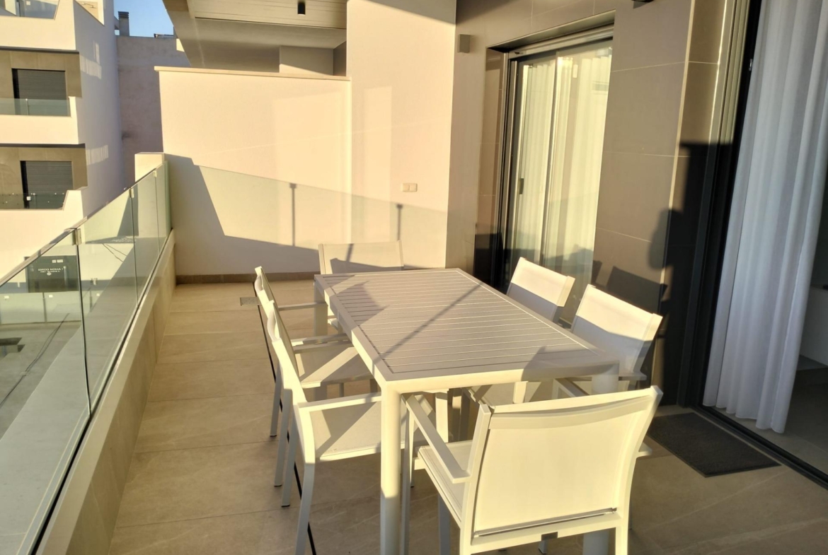 Modern balcony with a white outdoor dining table and matching chairs, glass railing along the edge, and a sliding glass door.