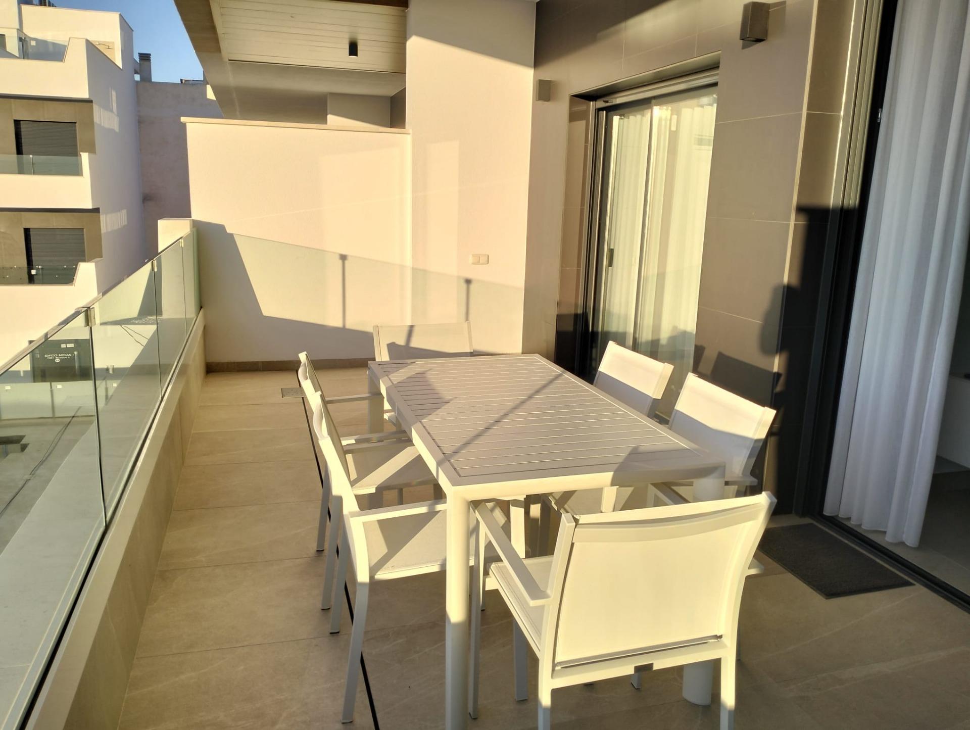 Modern balcony with a white outdoor dining table and matching chairs, glass railing along the edge, and a sliding glass door.