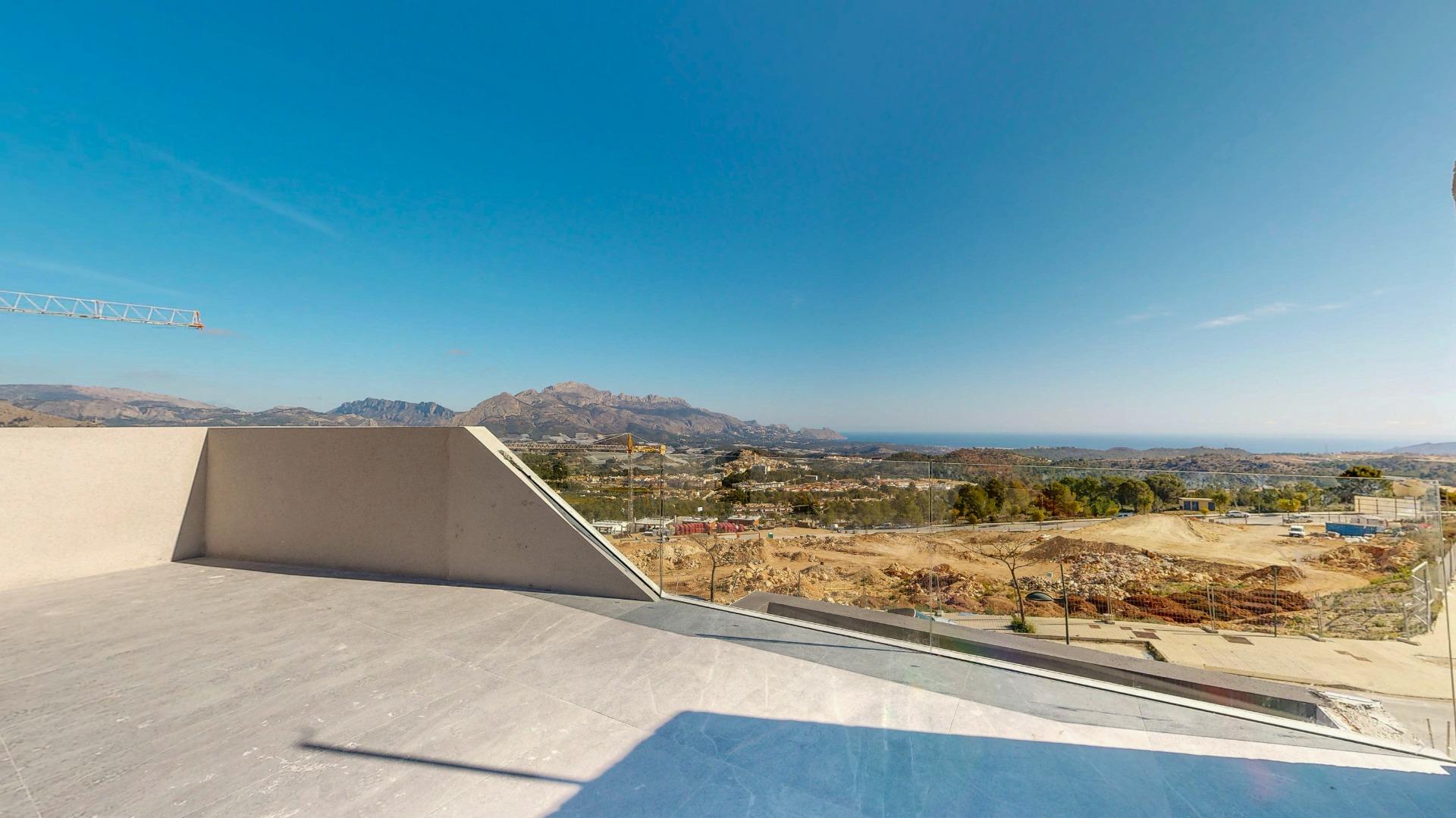 Construction site in the foreground with flat concrete and an angular beige wall; a crane appears on the left, with distant hills and the sea beyond under a clear blue sky.