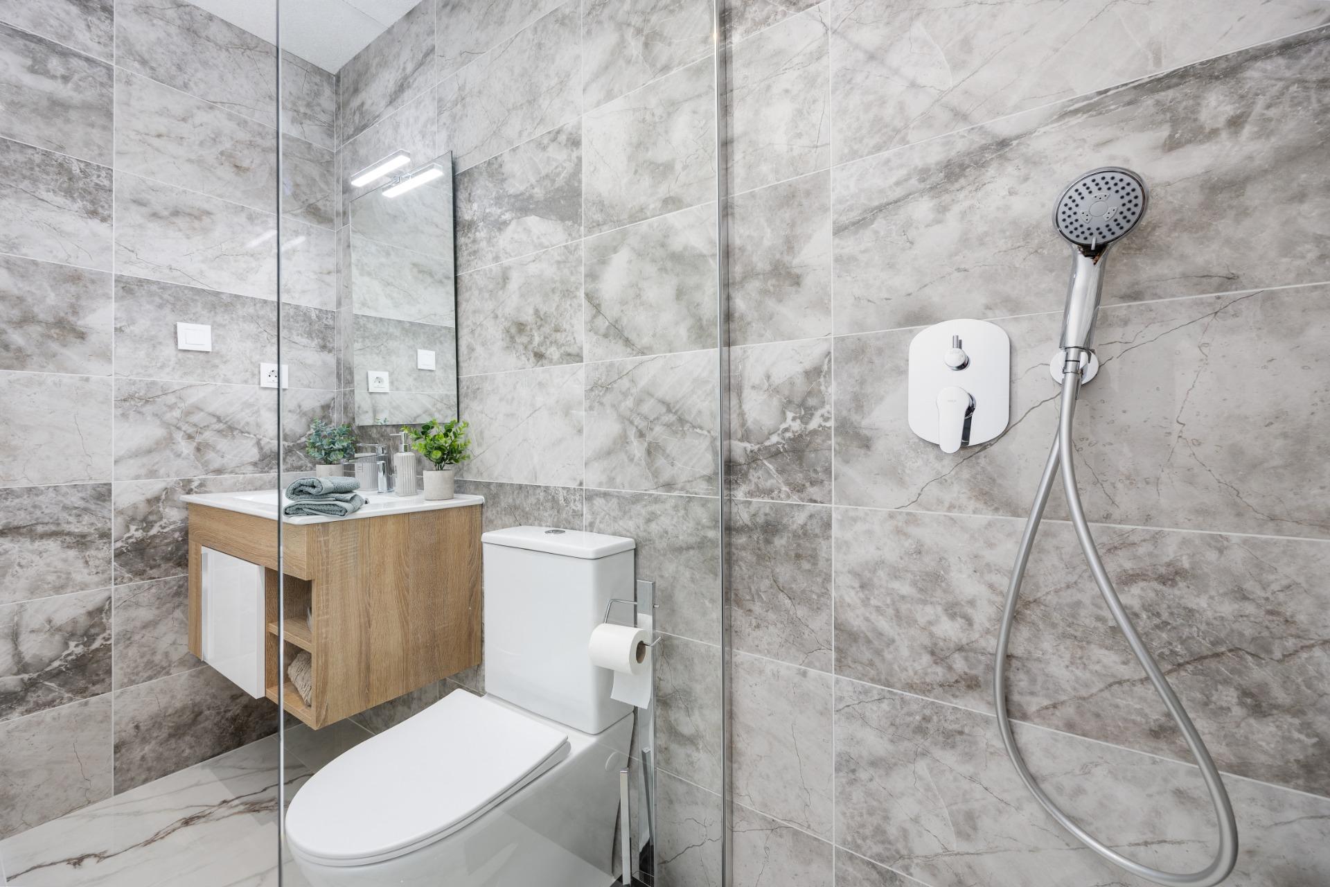 Modern bathroom with gray marble walls, glass shower partition, and a wooden vanity with towels and plants.