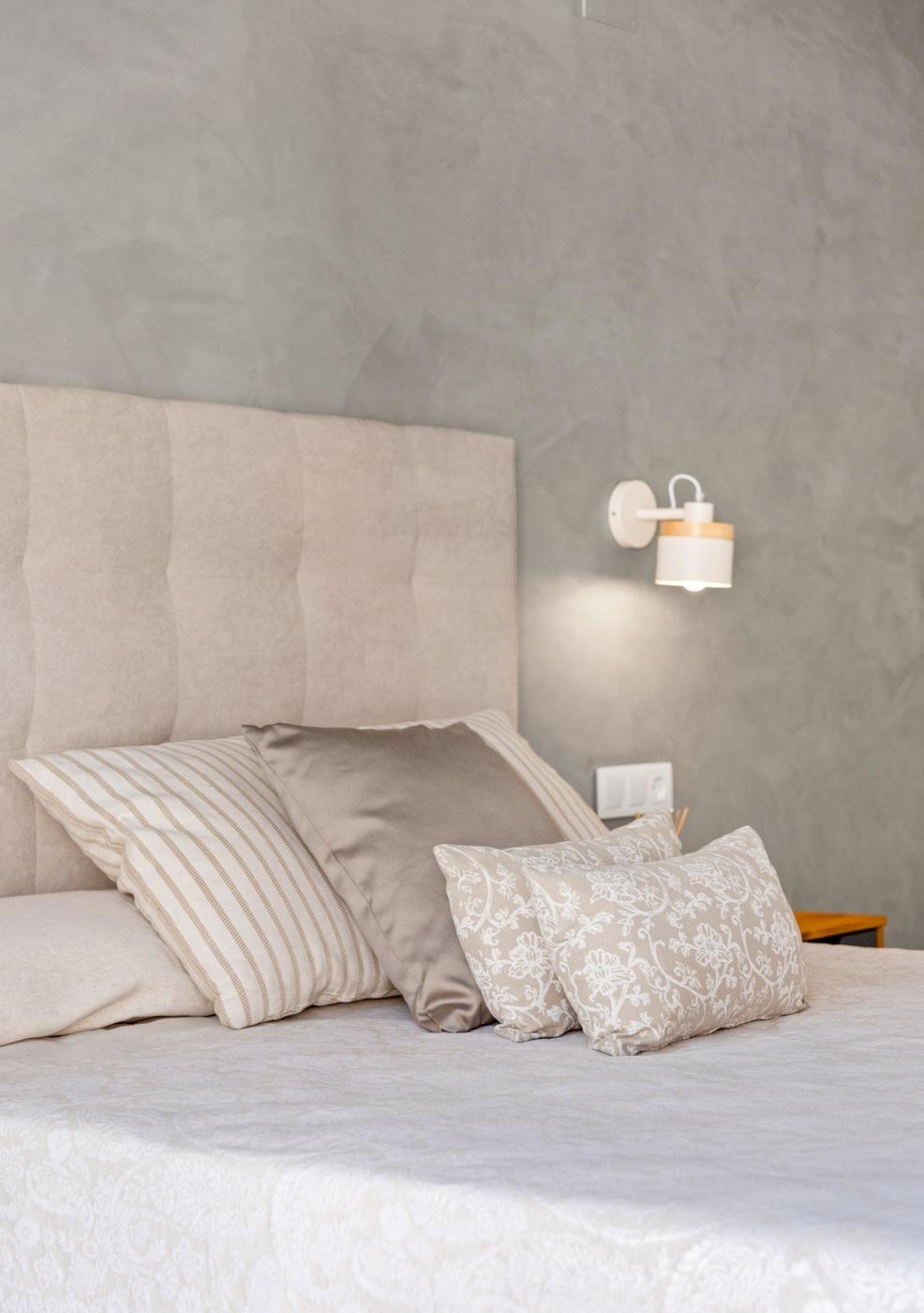 Bed with a tufted beige headboard and a row of neutral pillows against a gray textured wall with a wall-mounted lamp.