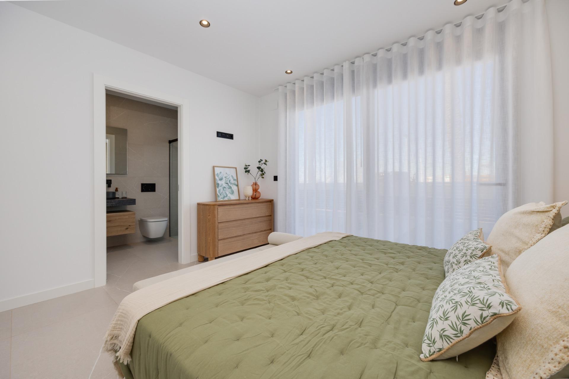 Bedroom with green quilted bed, white walls, sheer curtains, and light wood dresser; bathroom door open in background.