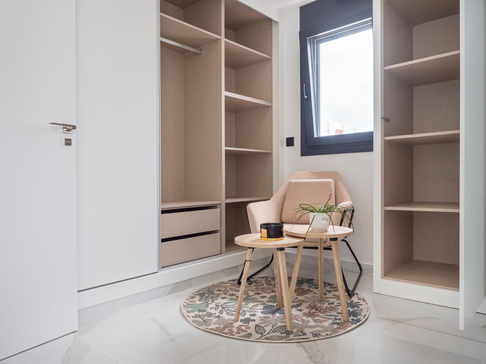 Walk-in closet with beige open shelves, a small round table and chair on a floral rug by a window, in daylight.