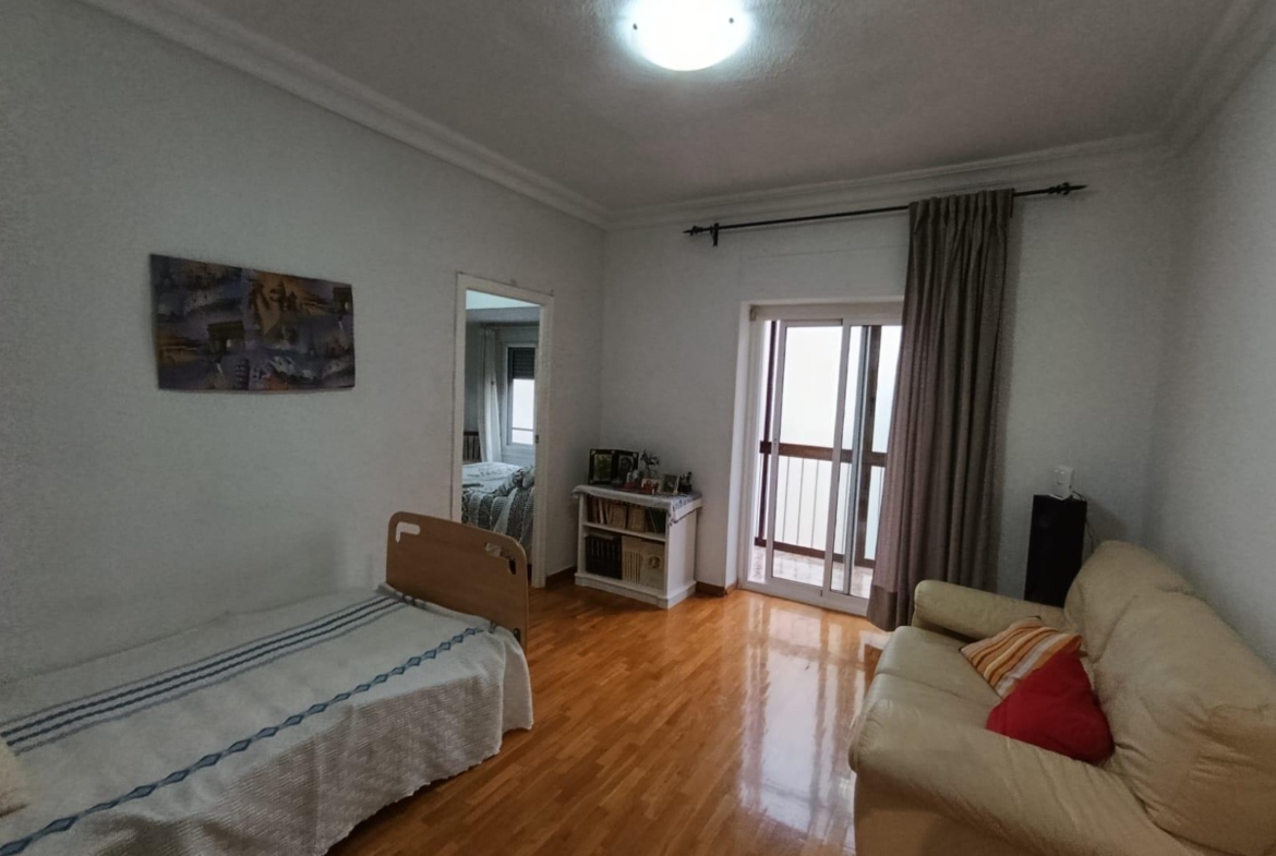 Living room with a beige sofa to the right, a single bed along the left wall, a white bookshelf and doorway to another room, and a glass balcony door with curtains on the far wall.