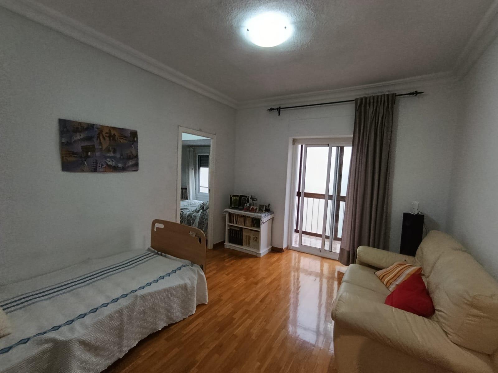 Living room with a beige sofa to the right, a single bed along the left wall, a white bookshelf and doorway to another room, and a glass balcony door with curtains on the far wall.