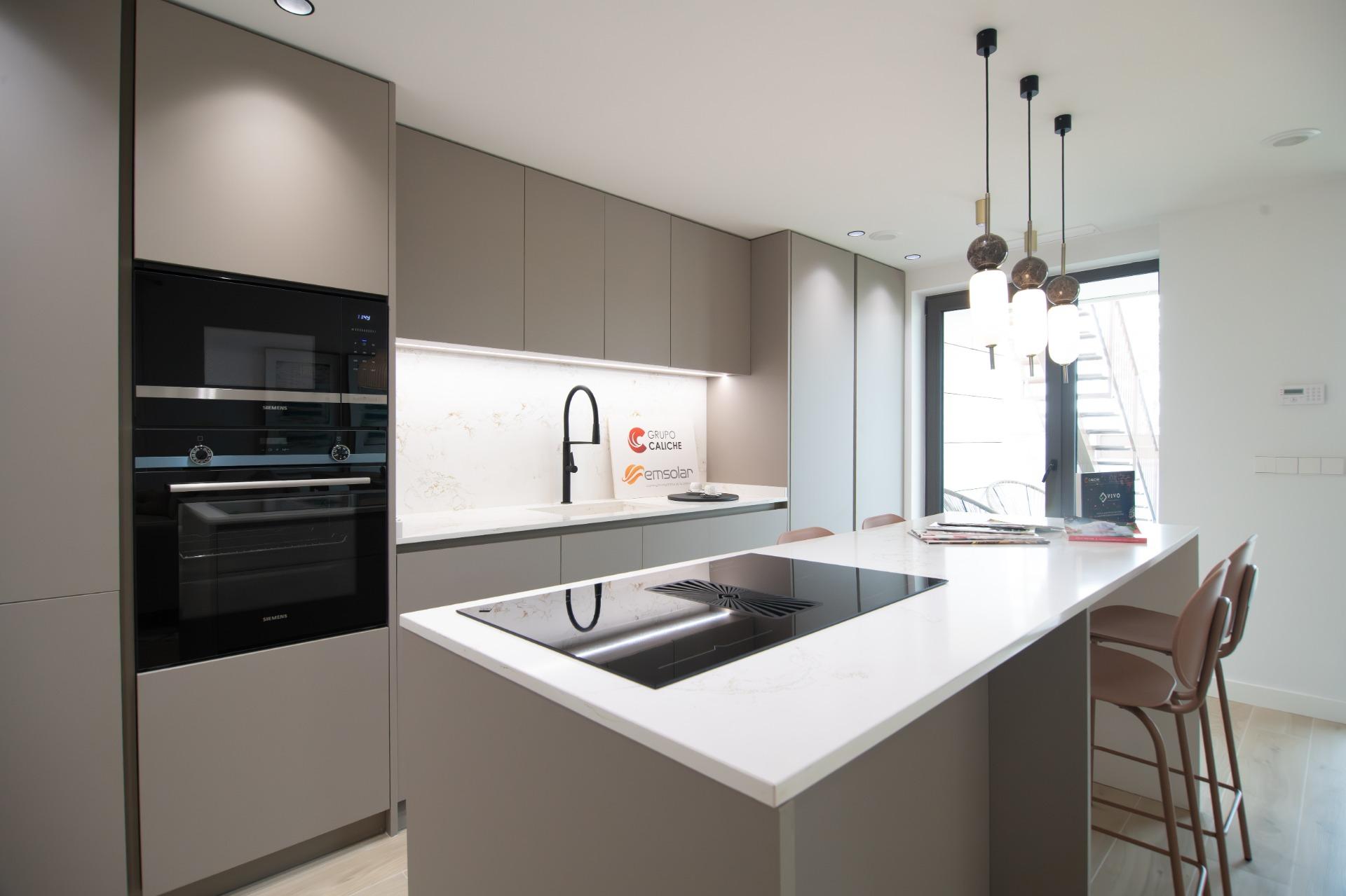 Modern kitchen with gray cabinetry, marble backsplash, and a large white island featuring a built-in cooktop and black faucet centerpiece; pendant lights overhead near a glass door.