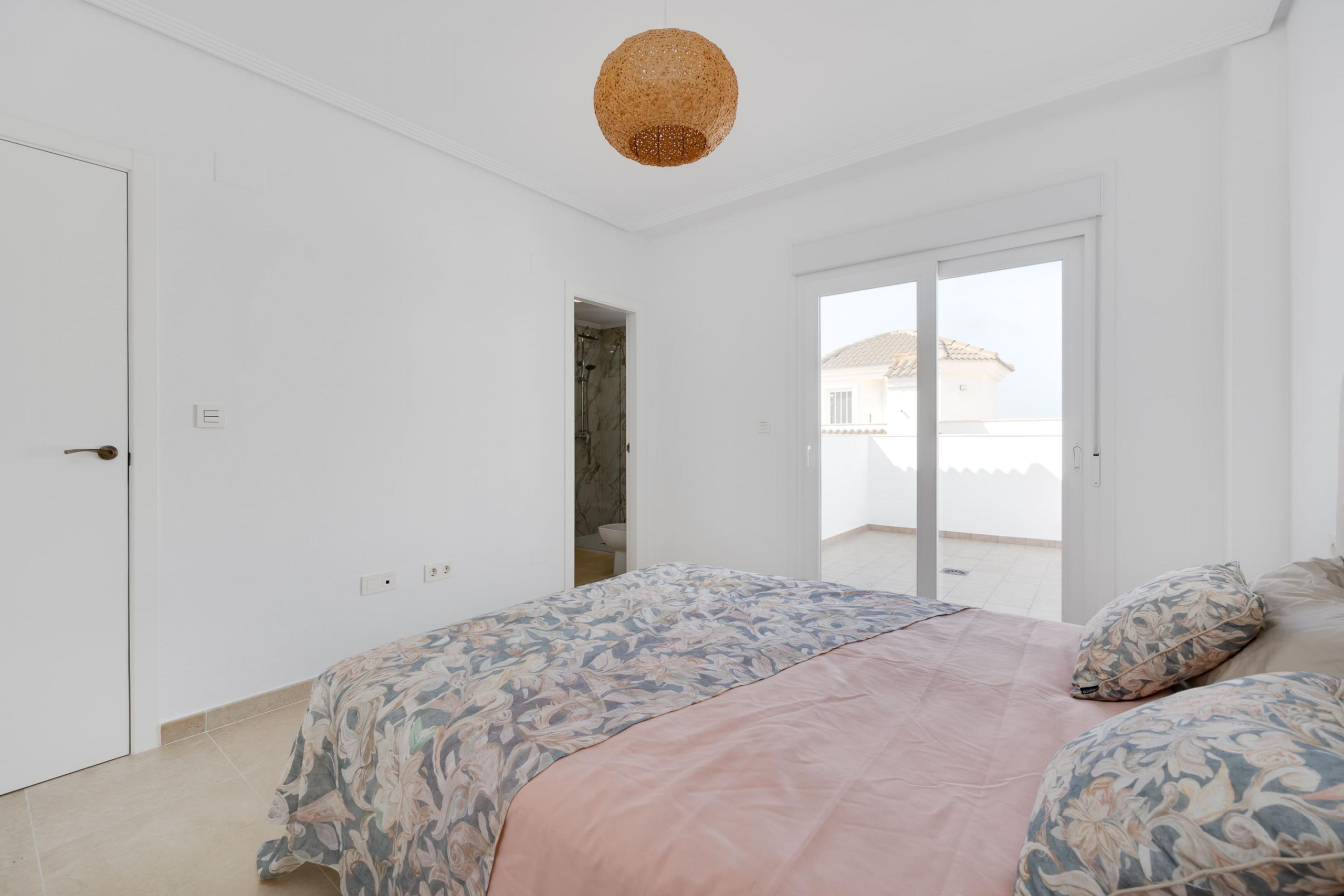 Bright white bedroom with floral quilted bedcovers, tiled floor, and sliding glass doors to a balcony.