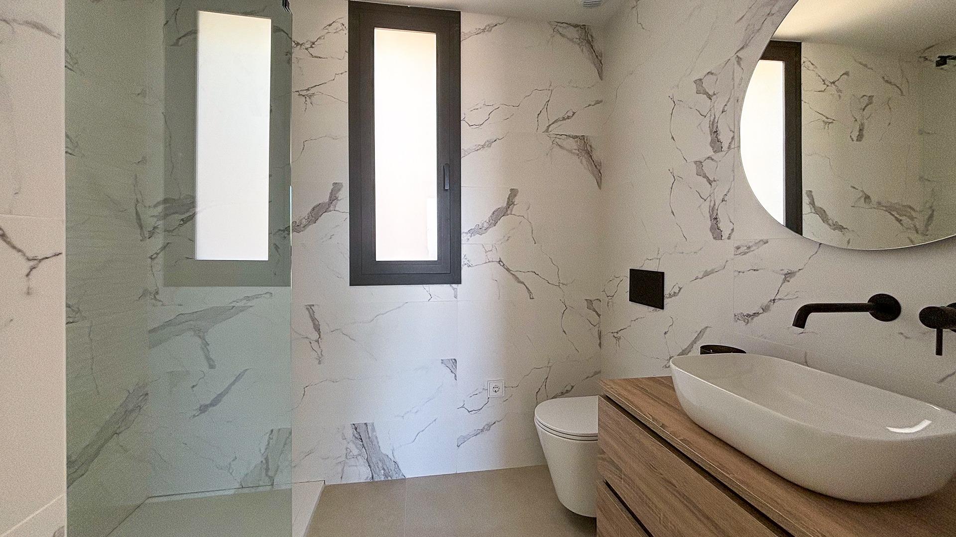 Modern marble-tiled bathroom with a dark-framed window, oval vessel sink on a wooden vanity, and black fixtures including faucet and wall controls.