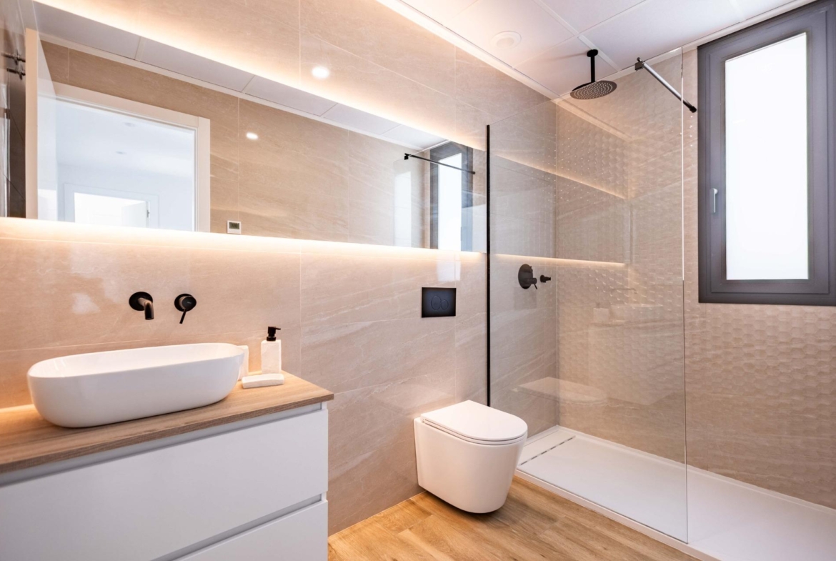 Modern bathroom with beige tiled walls, glass shower, black wall-mounted faucet, white vessel sink on a wood countertop, and a wall-hung toilet.
