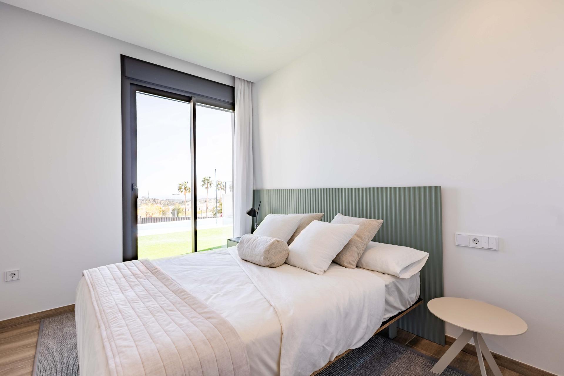 Bright modern bedroom with a low bed, white linens, beige pillows, and a green ribbed headboard beside a large glass door showing palm trees outside.