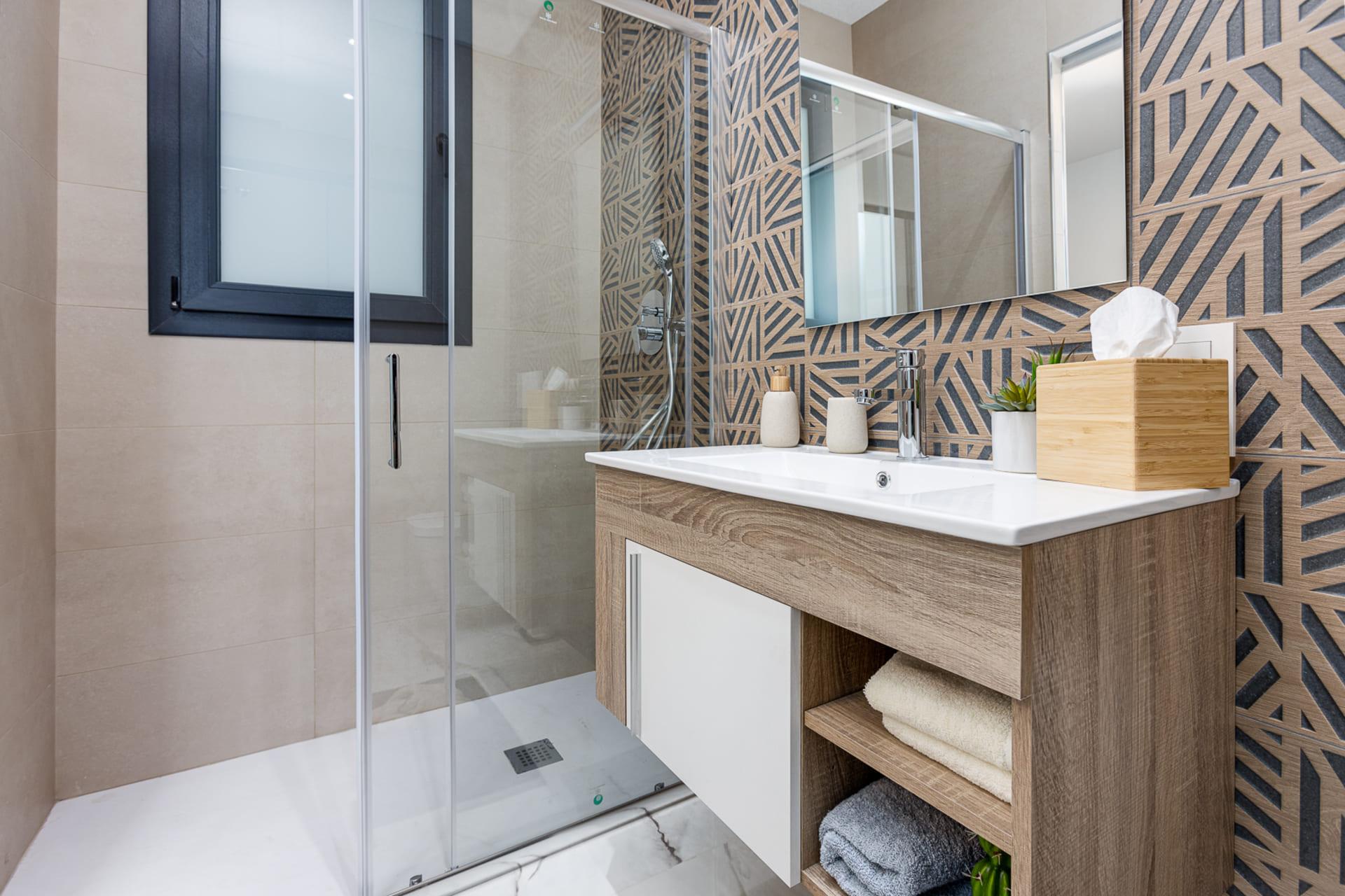 Modern bathroom featuring a glass shower enclosure, geometric patterned wall tiles, and a white sink with a wooden vanity. A tissue box, small plants, and neatly folded towels are visible on the counter and shelves.