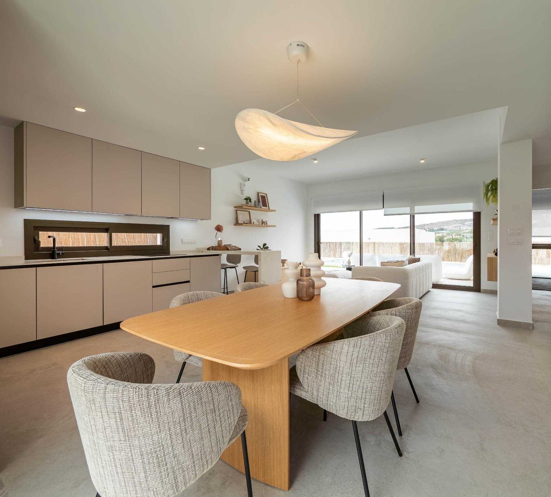 Open-plan kitchen and dining area with a light wood dining table, beige cabinets, and gray upholstered chairs under a sculptural pendant light.