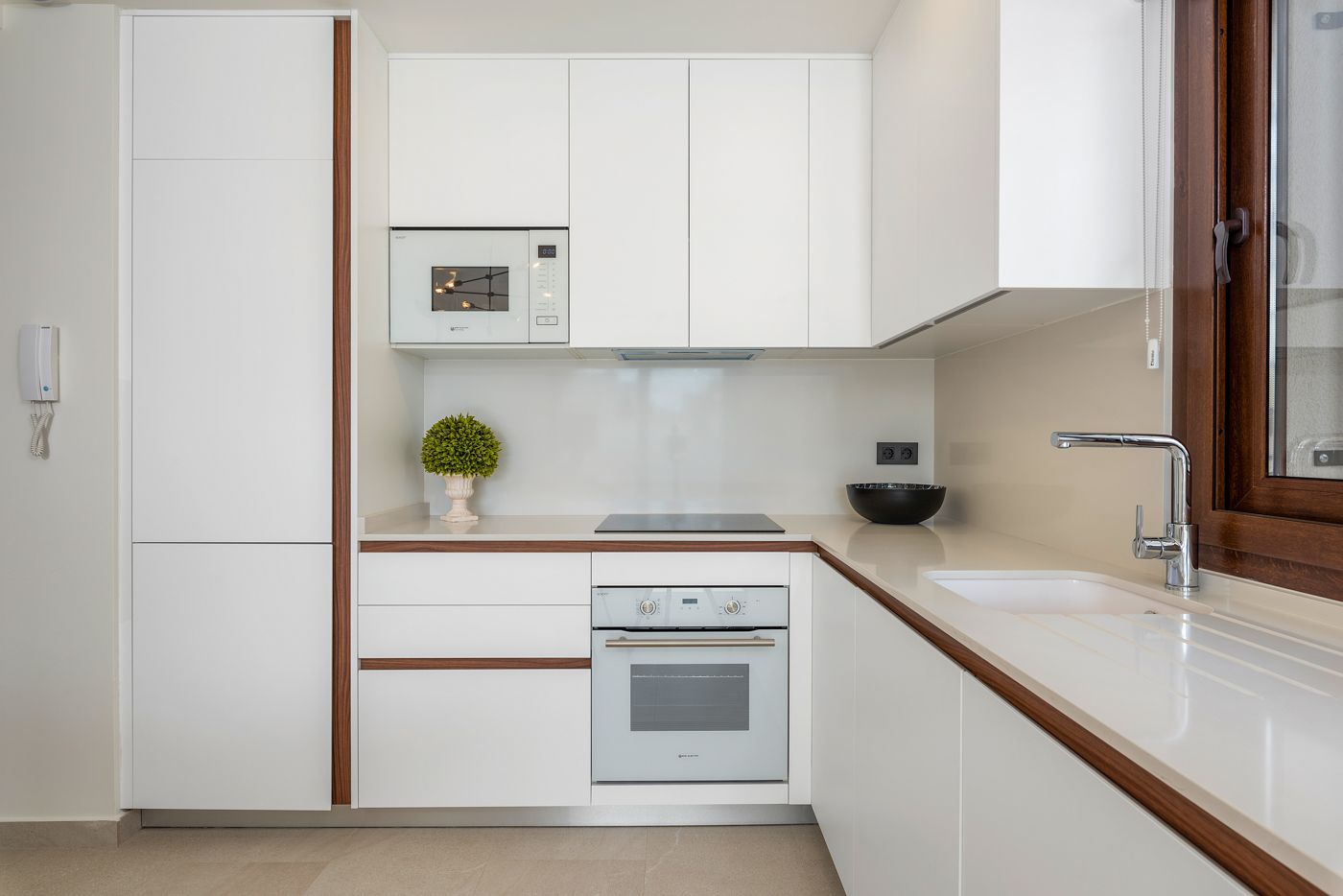 Modern white L-shaped kitchen with built-in oven, microwave, and sleek cabinetry; wooden trim accents and a large window over the sink