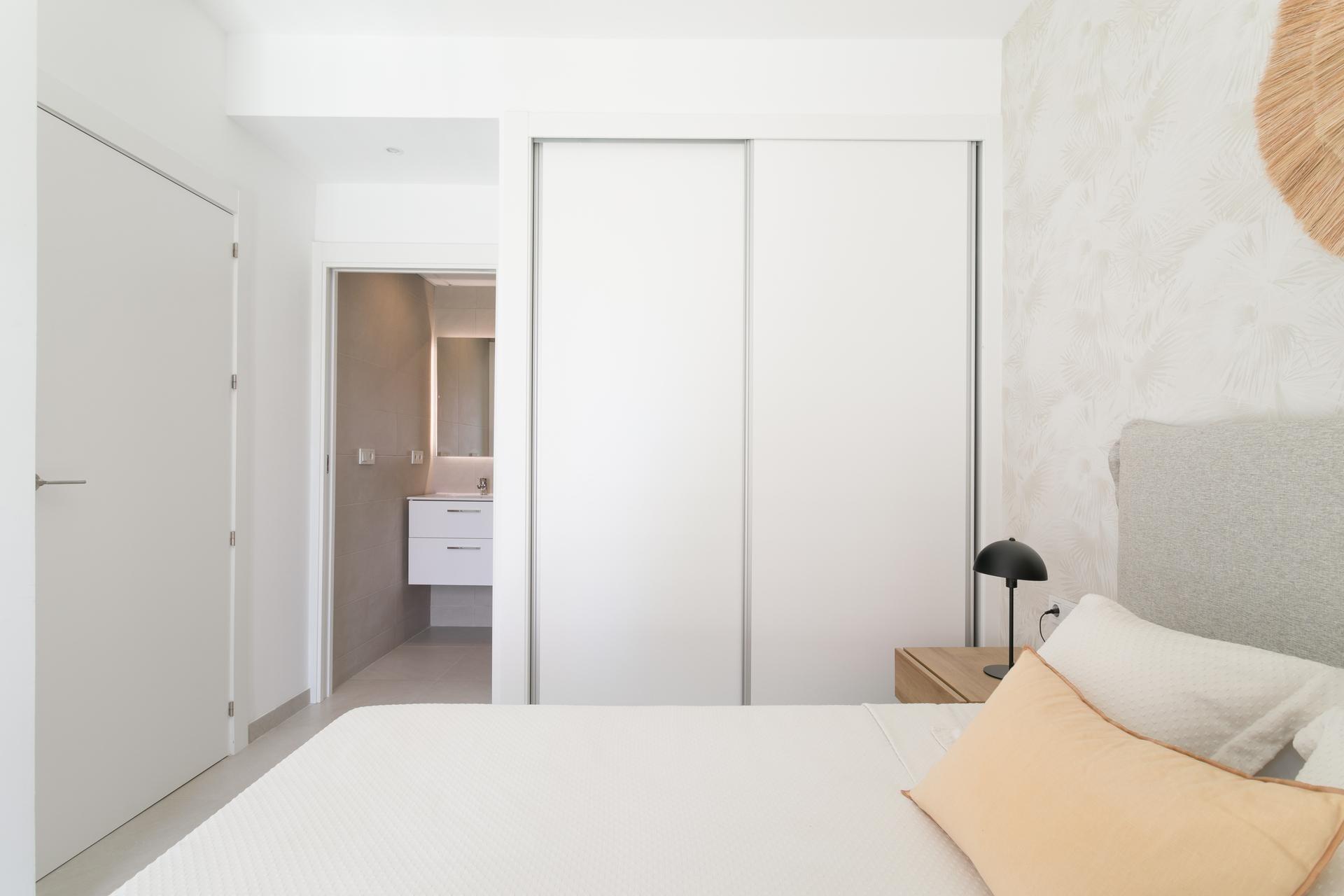 Bright minimalist bedroom with a large white sliding wardrobe, a bed with peach pillows, and a palm-patterned wall on the right.