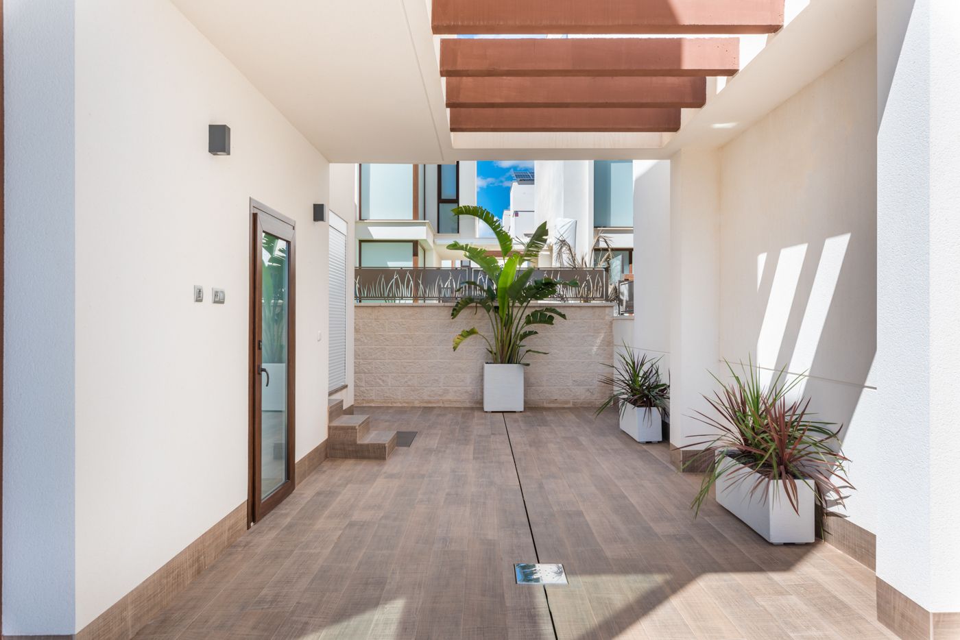 Modern outdoor corridor with white walls, wooden deck tiles, glass door on the left, and potted plants along the sides.