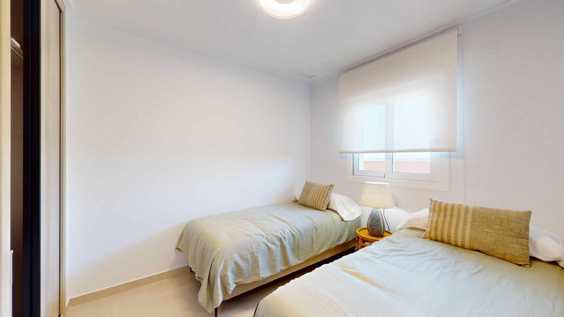 Twin bedroom with two single beds, beige comforters, and striped pillows against light walls; a nightstand and lamp between the beds.