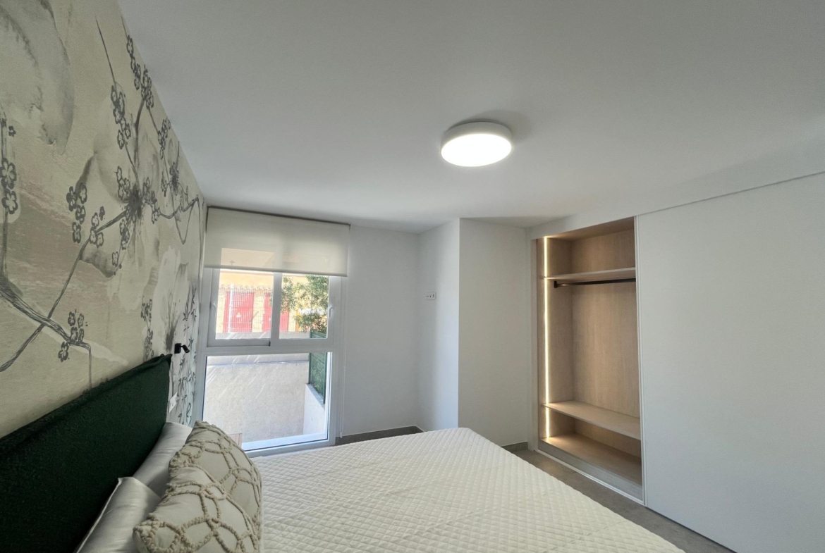 Bedroom with white walls and a floral mural on the left, a bed with a quilted white cover and decorative pillows, a window, and a built-in open wardrobe on the right.