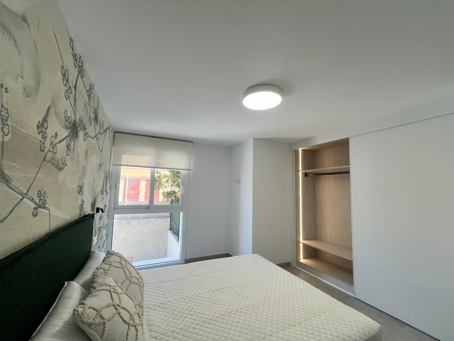 Bedroom with white walls and a floral mural on the left, a bed with a quilted white cover and decorative pillows, a window, and a built-in open wardrobe on the right.