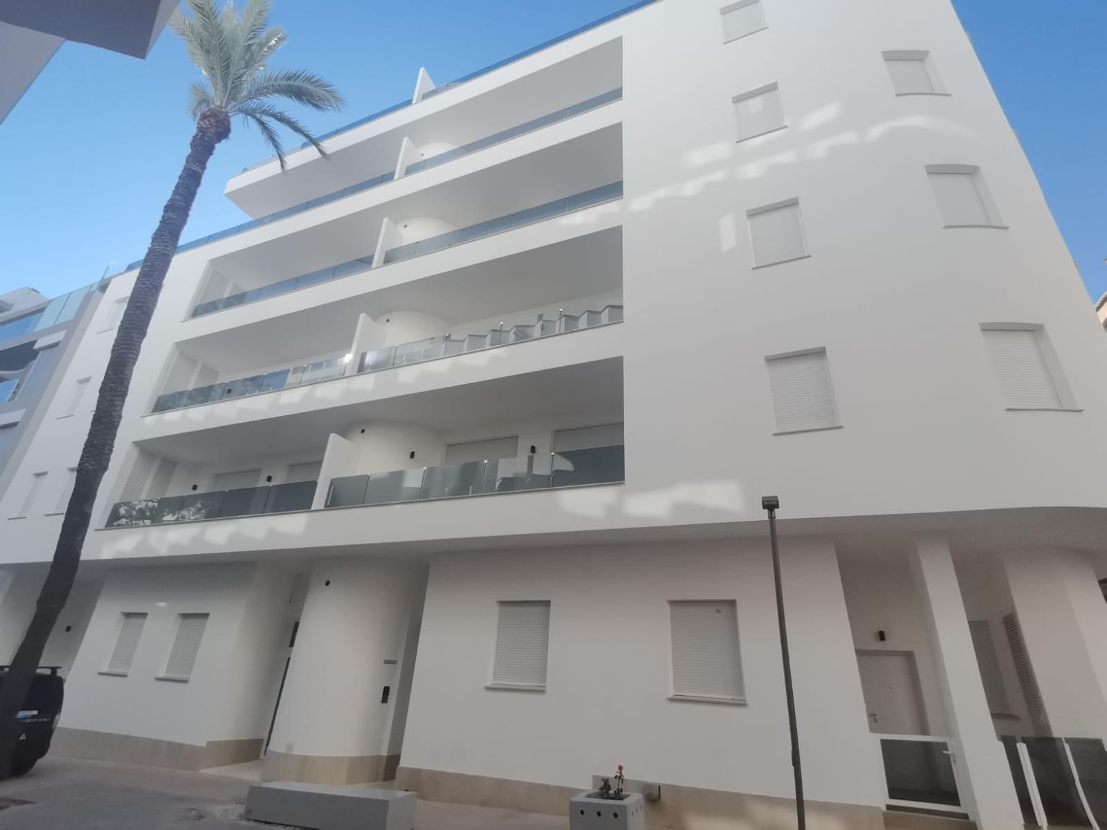 White modern apartment building with several balconies, glass railings, and a tall palm tree in the foreground against a blue sky.