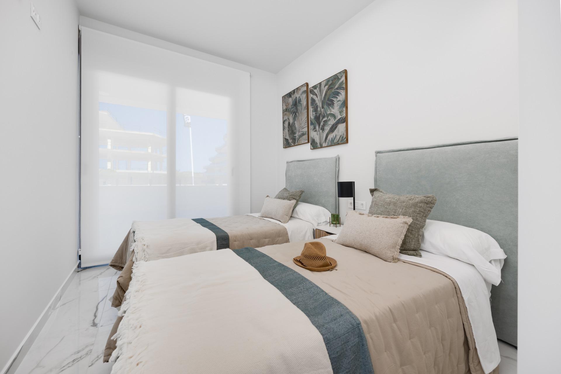 Twin bedroom with two single beds, beige duvets with teal runners, gray headboards, and a nightstand between them. Large window with sheer blinds visible outside.