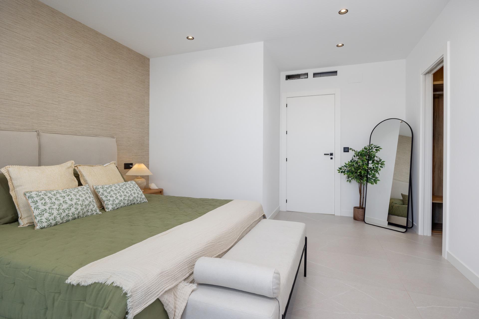 Bedroom with a beige textured accent wall behind a light gray headboard and green quilted bedspread with multiple decorative pillows. a bedside lamp on a wooden nightstand is lit.