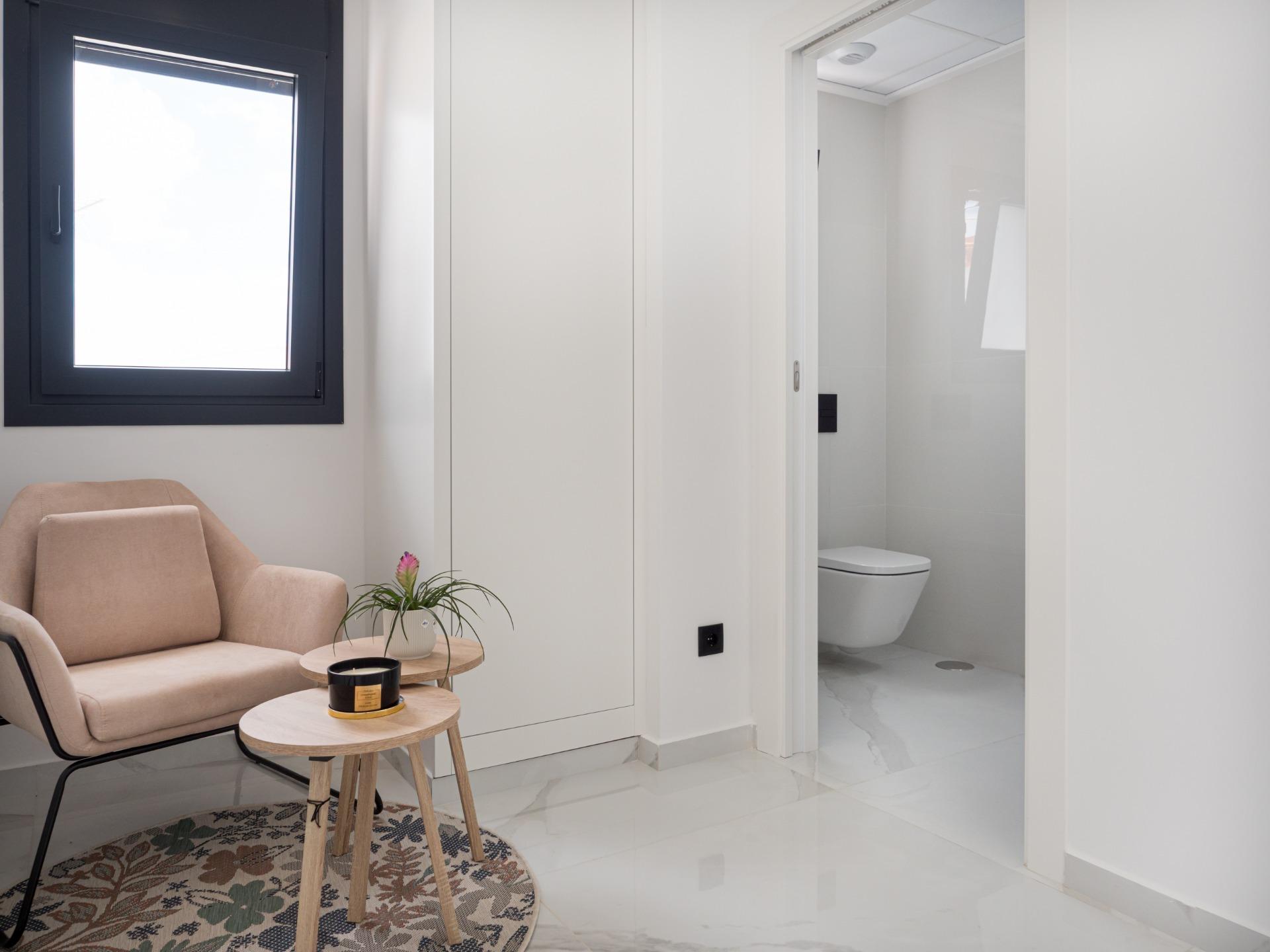 Modern living area with beige armchairs, two round wooden tables, a potted plant, and a floral rug; doorway to a white-tiled bathroom on the right