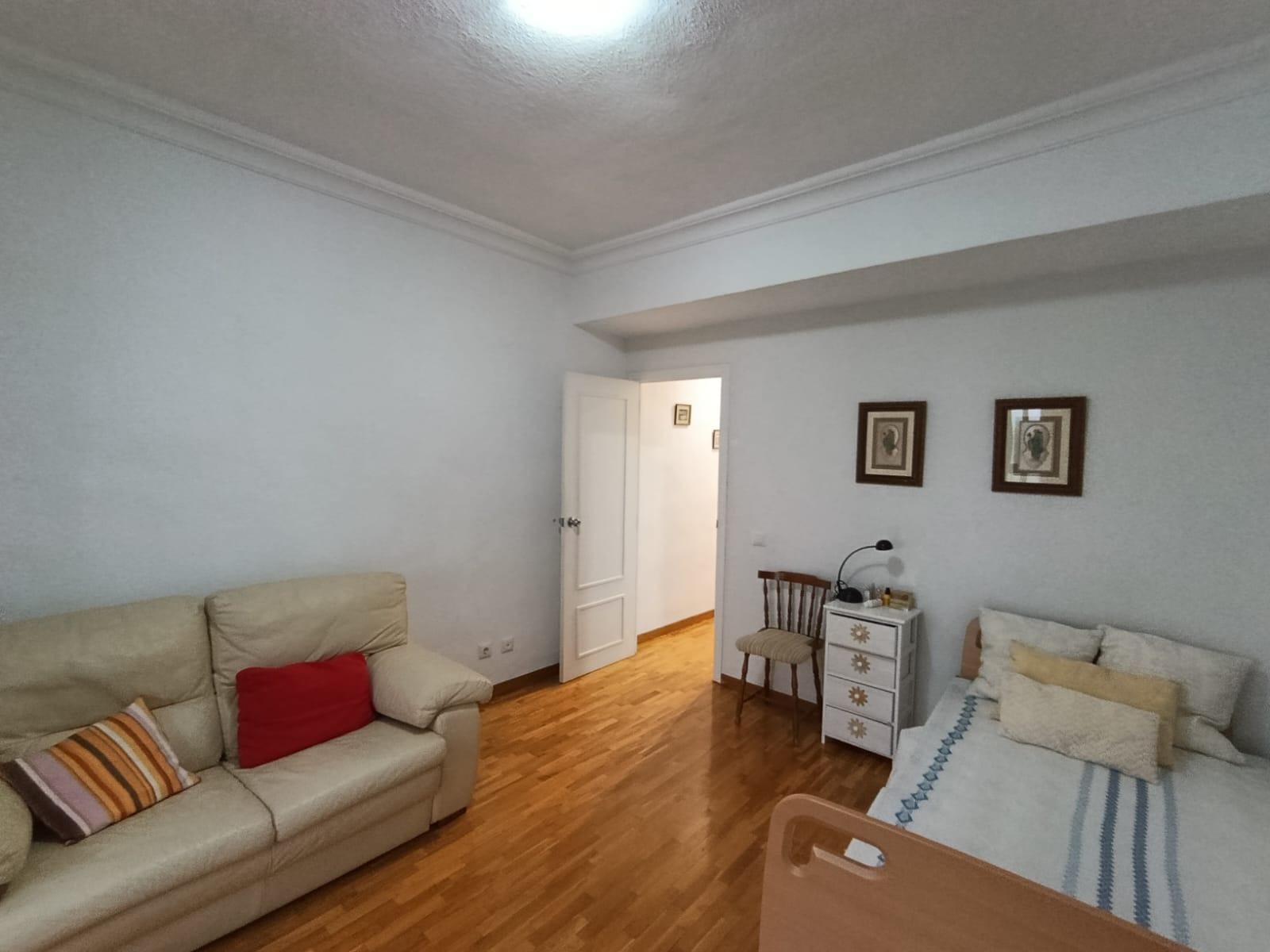 Living room with beige sofa, red cushion, and striped pillows facing a bed along the opposite wall; wooden floor and soft lighting.