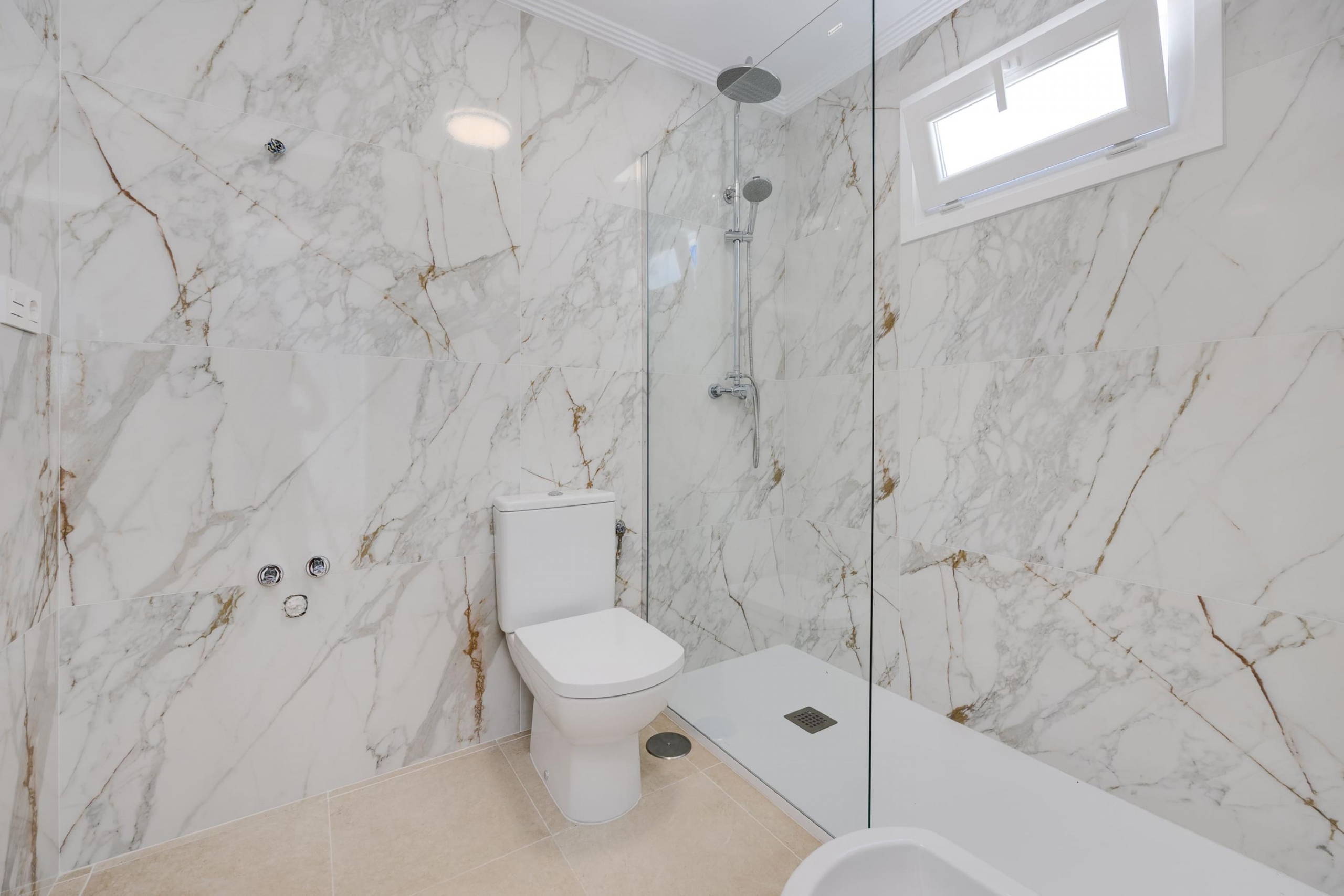 Marble-tiled bathroom with a glass shower enclosure, chrome showerhead, and a white toilet under a bright window.