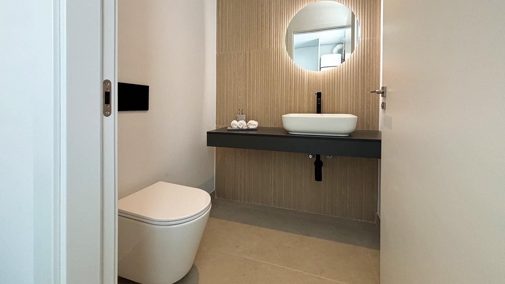 Modern bathroom with a wall-mounted dark vanity, white vessel sink, circular backlit mirror, and beige textured wall tiles.