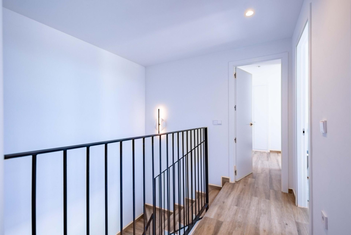 Bright upstairs hallway with a black metal railing, light wood floor, and an open door to a bright room outside the frame.