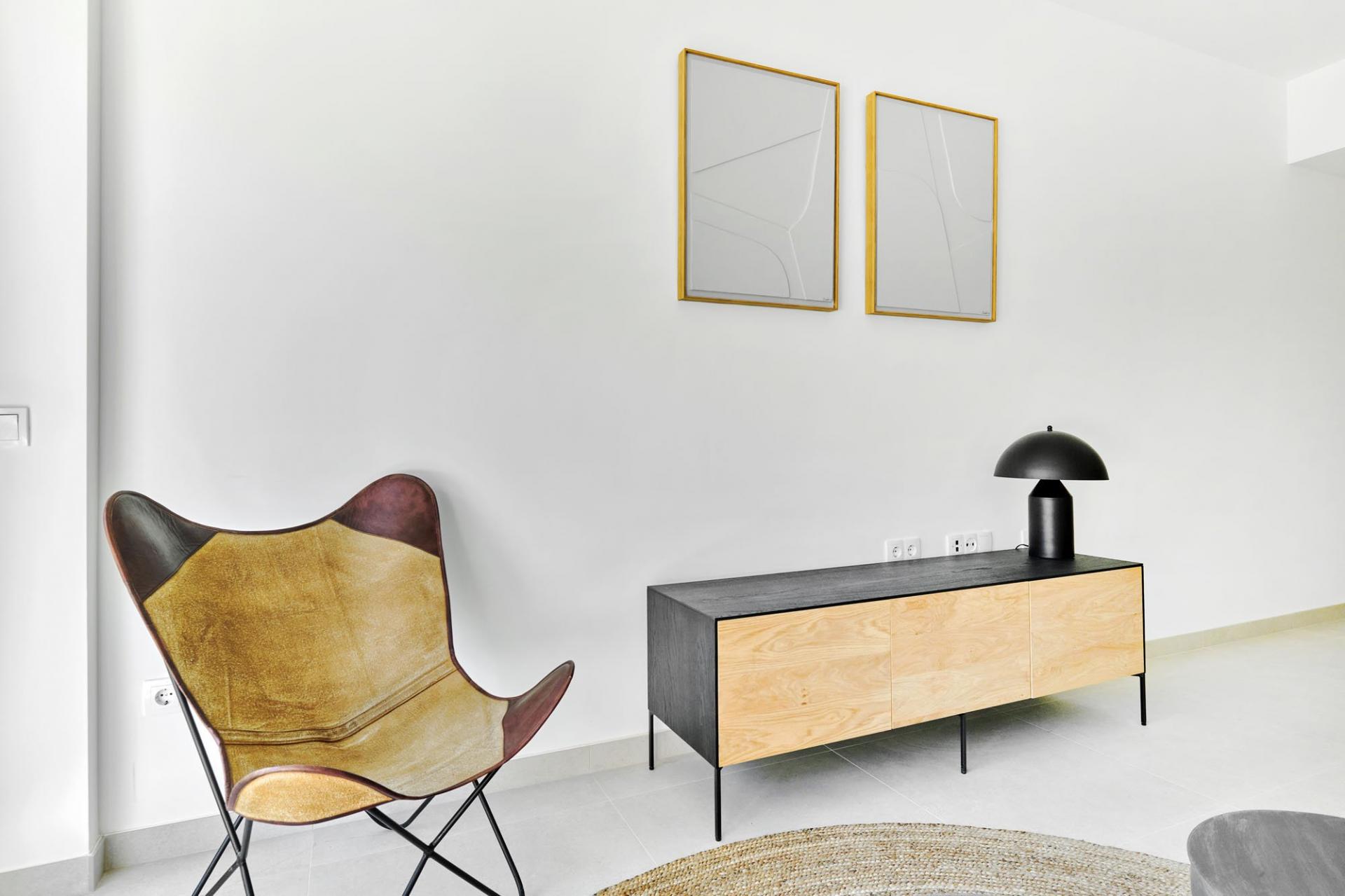 Mid-century butterfly chair with worn brown leather beside a two-tone wooden sideboard against a white wall with two square gold-framed abstract prints and a black lamp atop the cabinet.
