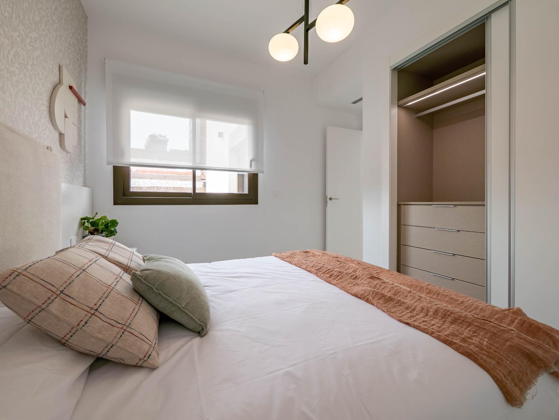 Modern bedroom with a made bed, plaid pillows, and an orange throw; window to the left and an open closet to the right.