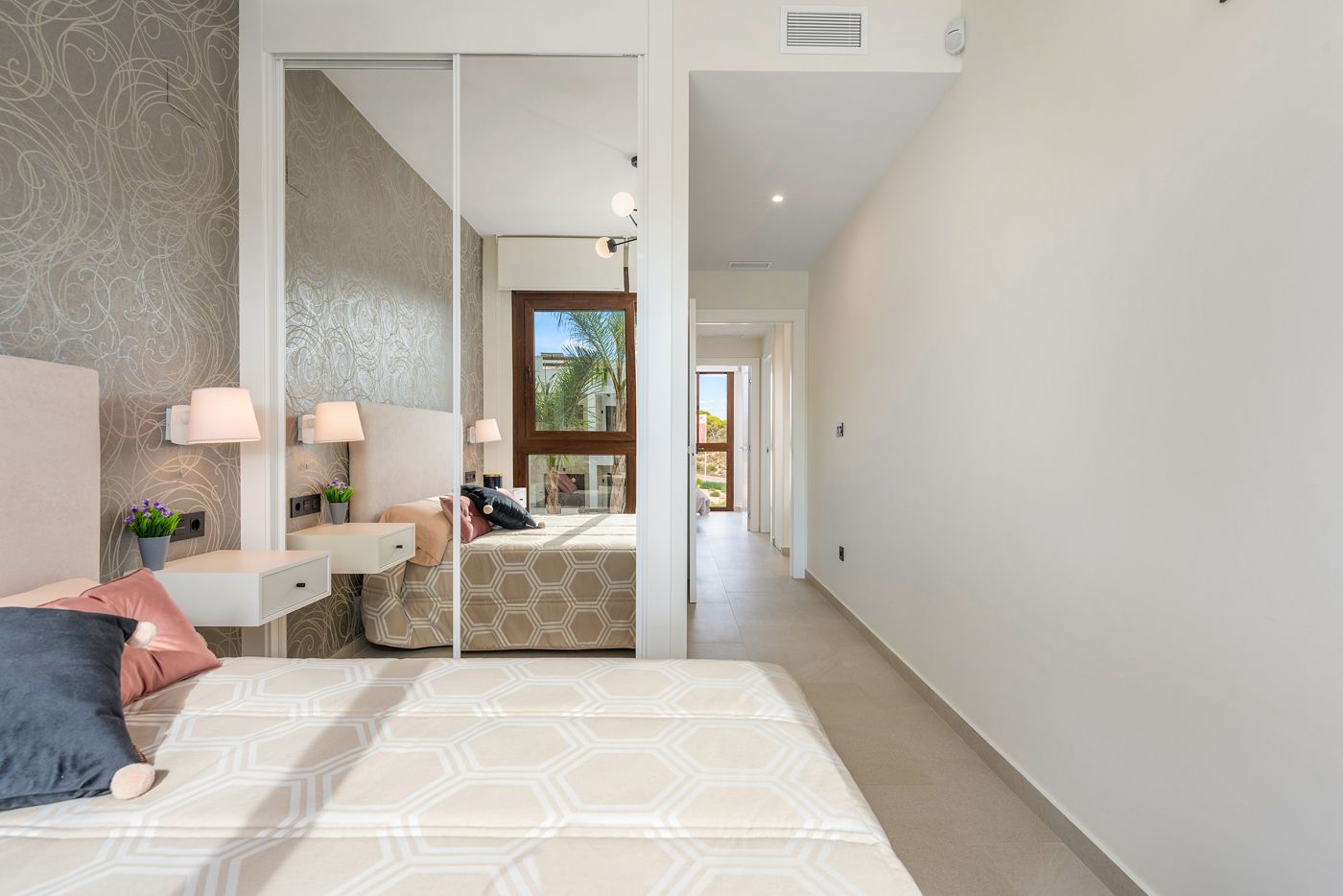 Bright modern hotel bedroom with a mirrored sliding wardrobe, two white bedside tables, and a bed with a beige hex pattern bedspread.