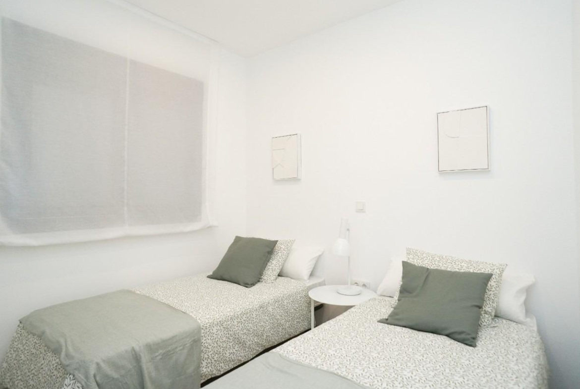 Two single beds with patterned linens and green pillows, a shared white nightstand with a lamp between them in a bright white room