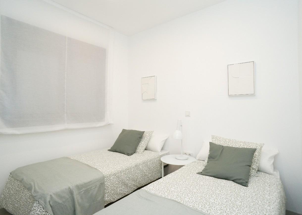 Two single beds with patterned linens and green pillows, a shared white nightstand with a lamp between them in a bright white room