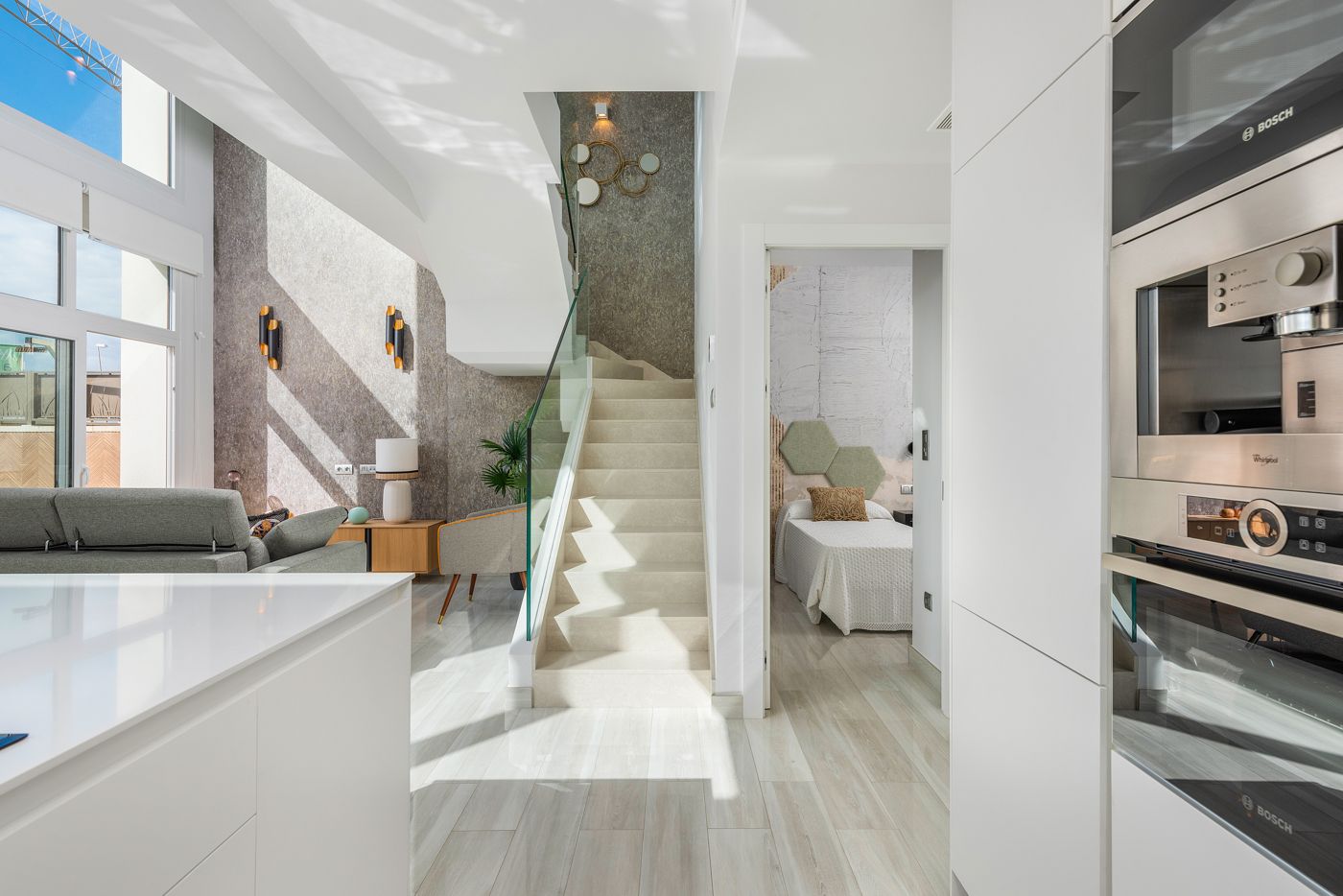 Bright, modern open-plan living space with a white kitchen island, gray sofa area, glass stair railing, and a doorway to a bedroom; built-in Bosch oven visible on the right.