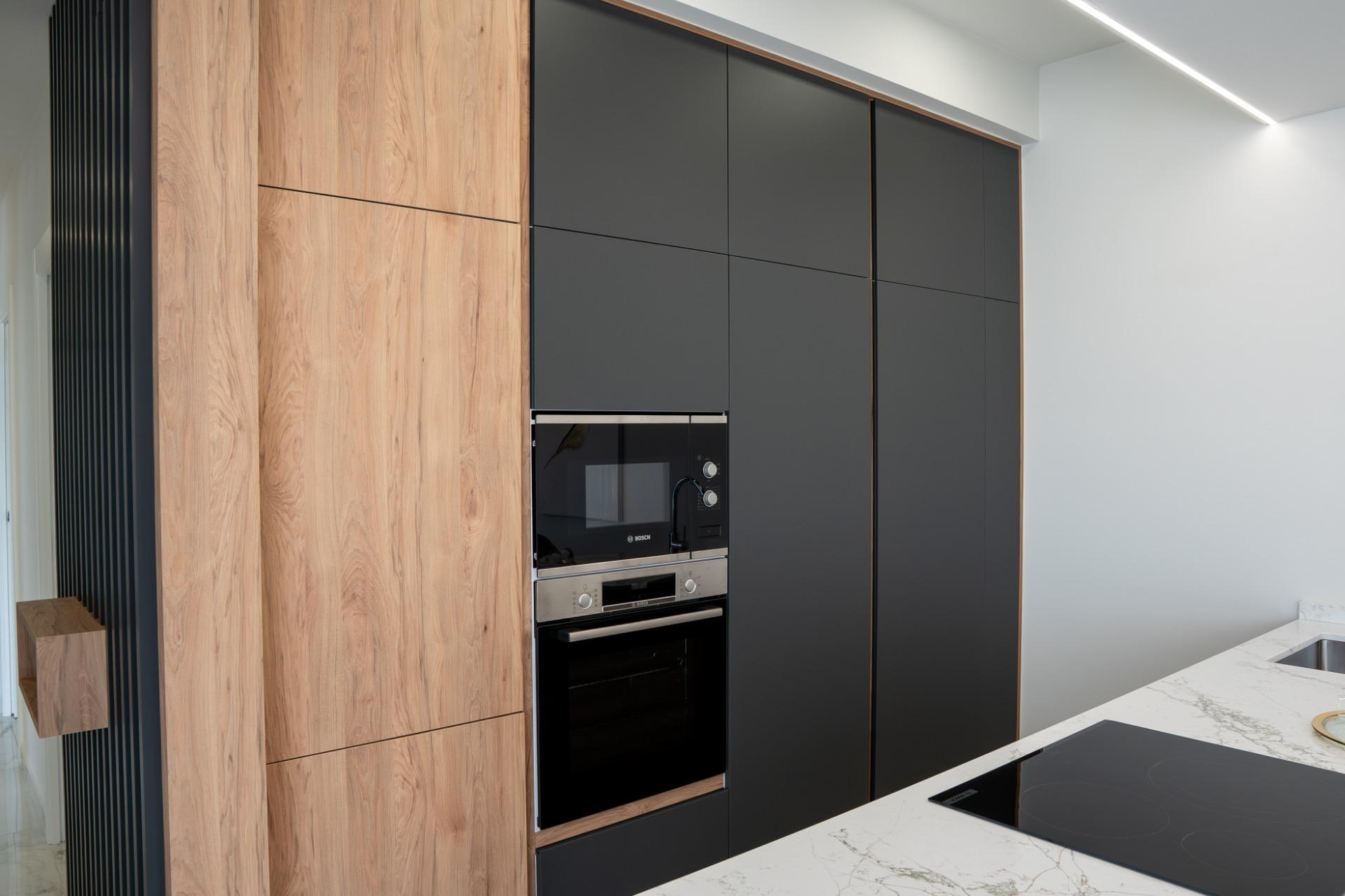 Modern kitchen with tall wood cabinets, matte dark-gray panels, and built-in Bosch oven and microwave set into the cabinetry, marble countertop visible in foreground.