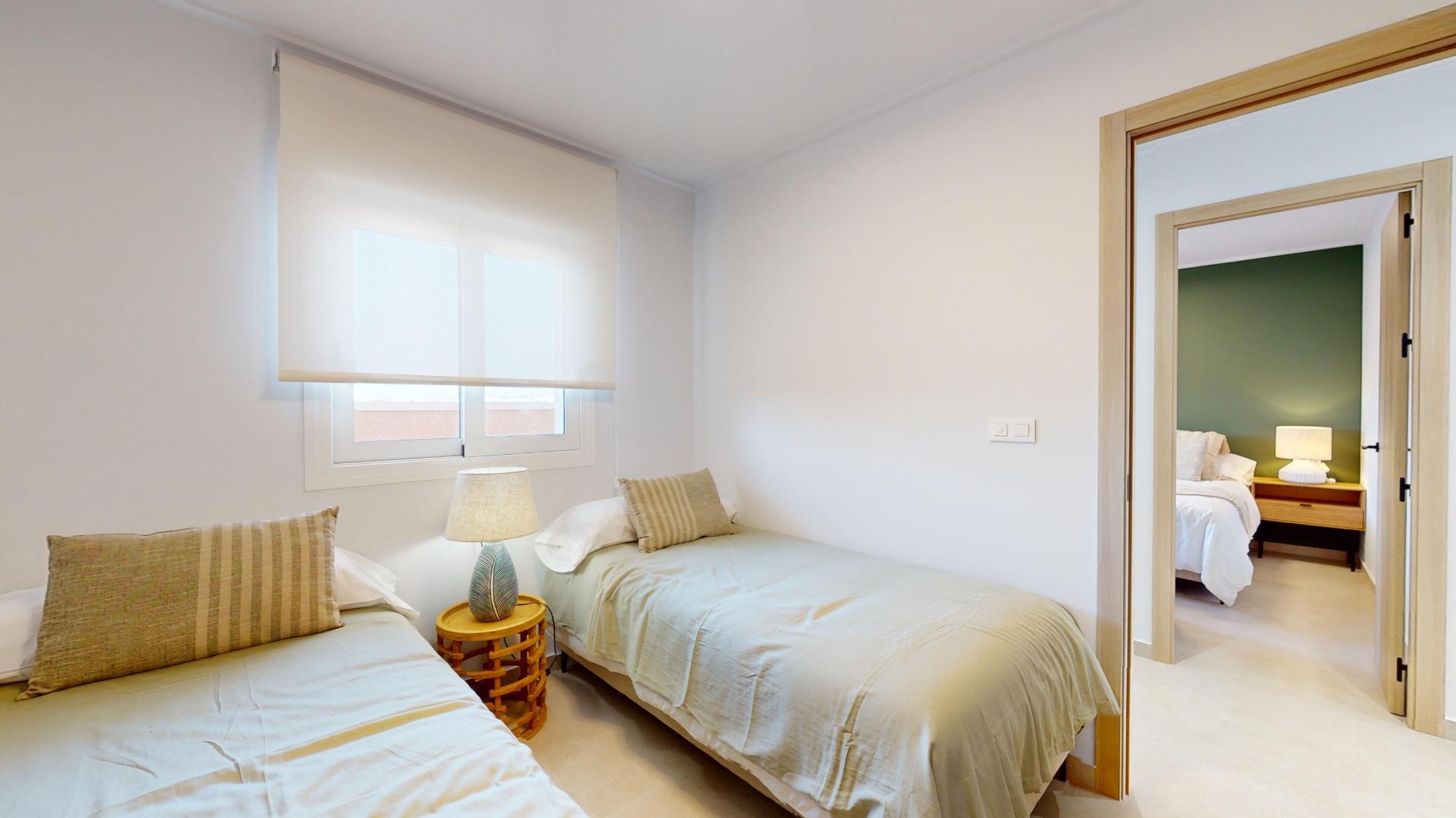 Twin bedroom with two single beds, beige bedding and striped pillows, a small lamp on a wooden table between them, and a window with white shades; open doorway shows a second room with a green accent wall.