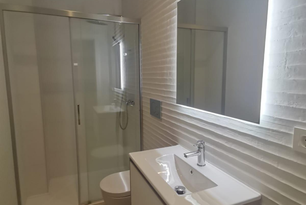 Modern bathroom with glass-enclosed shower, wall-mounted white sink vanity, and backlit rectangular mirror on a textured white wall.