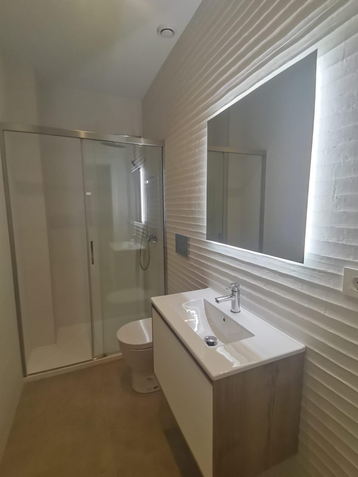 Modern bathroom with glass-enclosed shower, wall-mounted white sink vanity, and backlit rectangular mirror on a textured white wall.