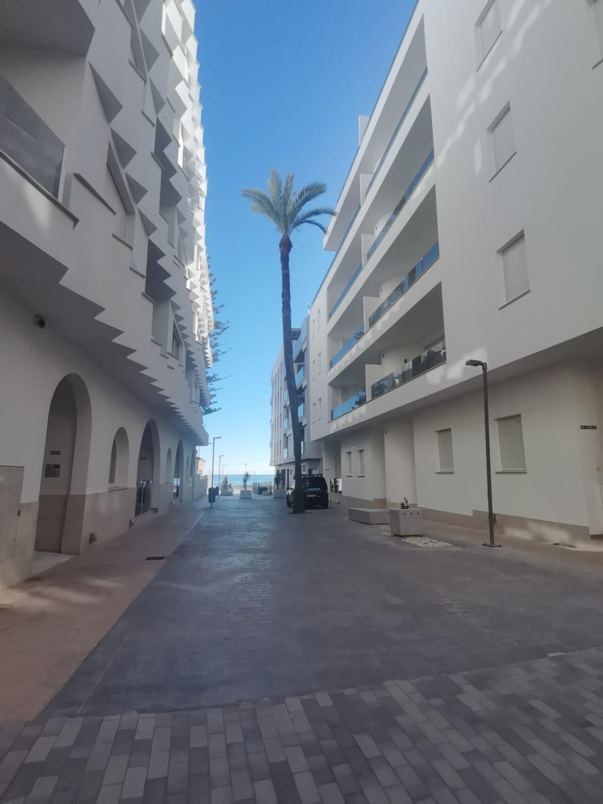 Narrow white street between modern apartment buildings, a tall palm tree in the center and blue sky, leading to the sea in the distance.
