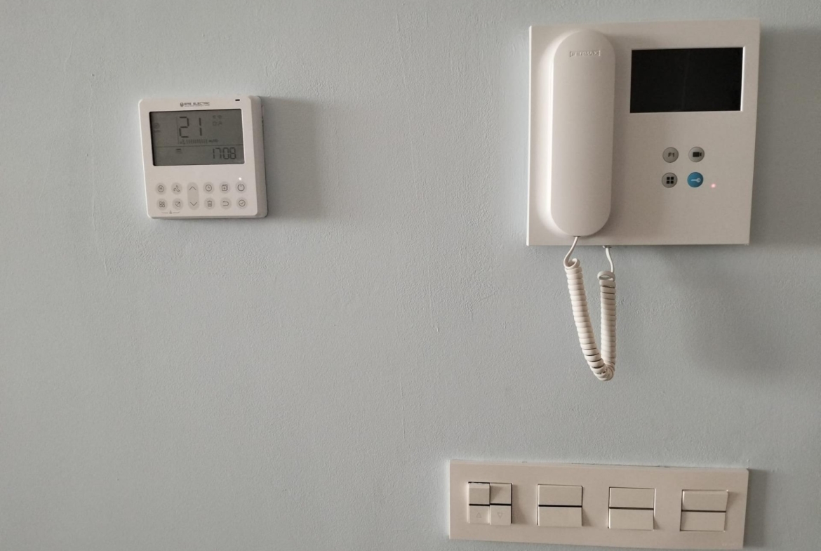 Wall thermostat on the left and a wall-mounted intercom with handset and small screen on the right, all mounted on a light gray wall, with a beige switch panel below.