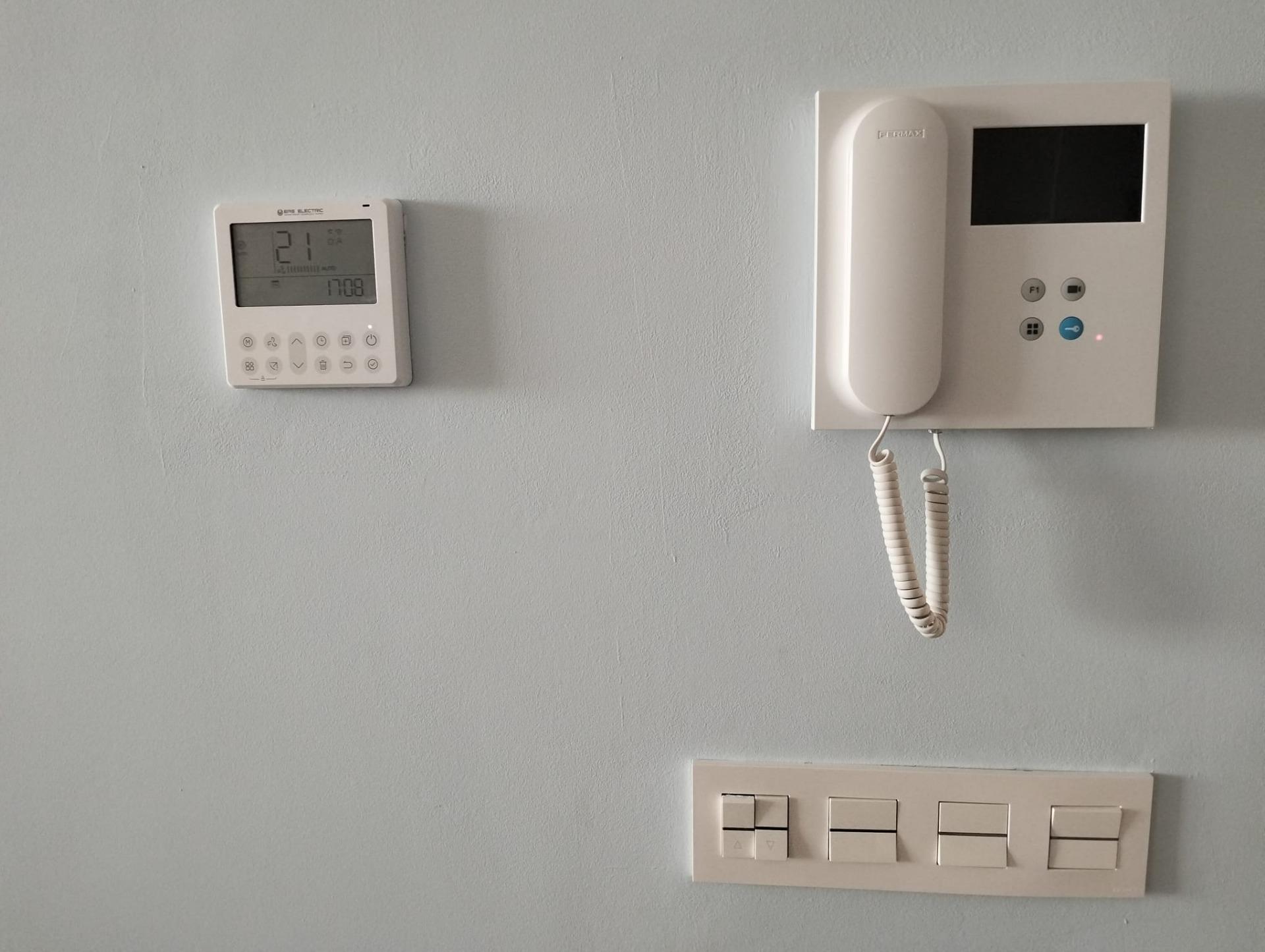 Wall thermostat on the left and a wall-mounted intercom with handset and small screen on the right, all mounted on a light gray wall, with a beige switch panel below.