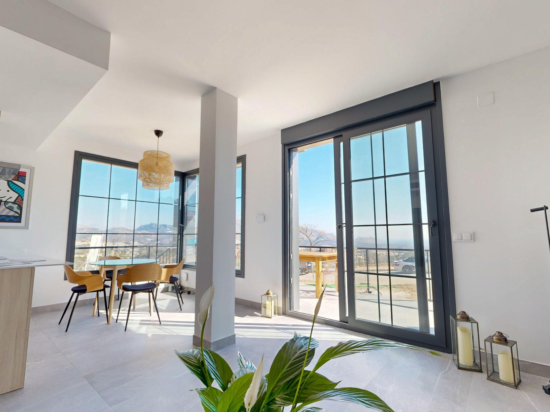 Bright open-plan dining area with large gray-framed glass doors, a round glass table, and wooden chairs; sunny mountains outside visible through windows.