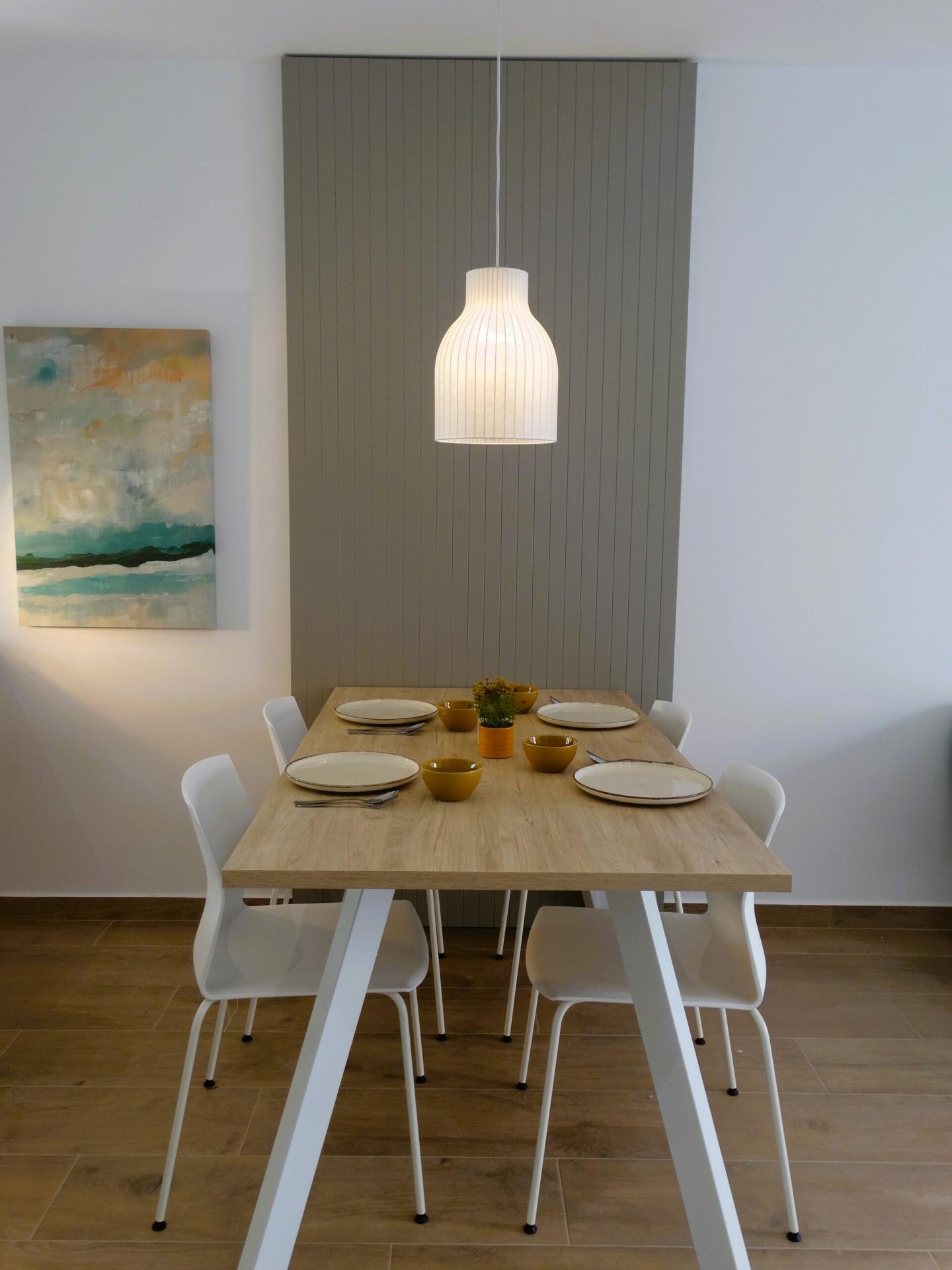 Dining room with a light wood table set for six, white chairs, yellow bowls and plates, and a white pendant lamp above; gray panel wall in back.