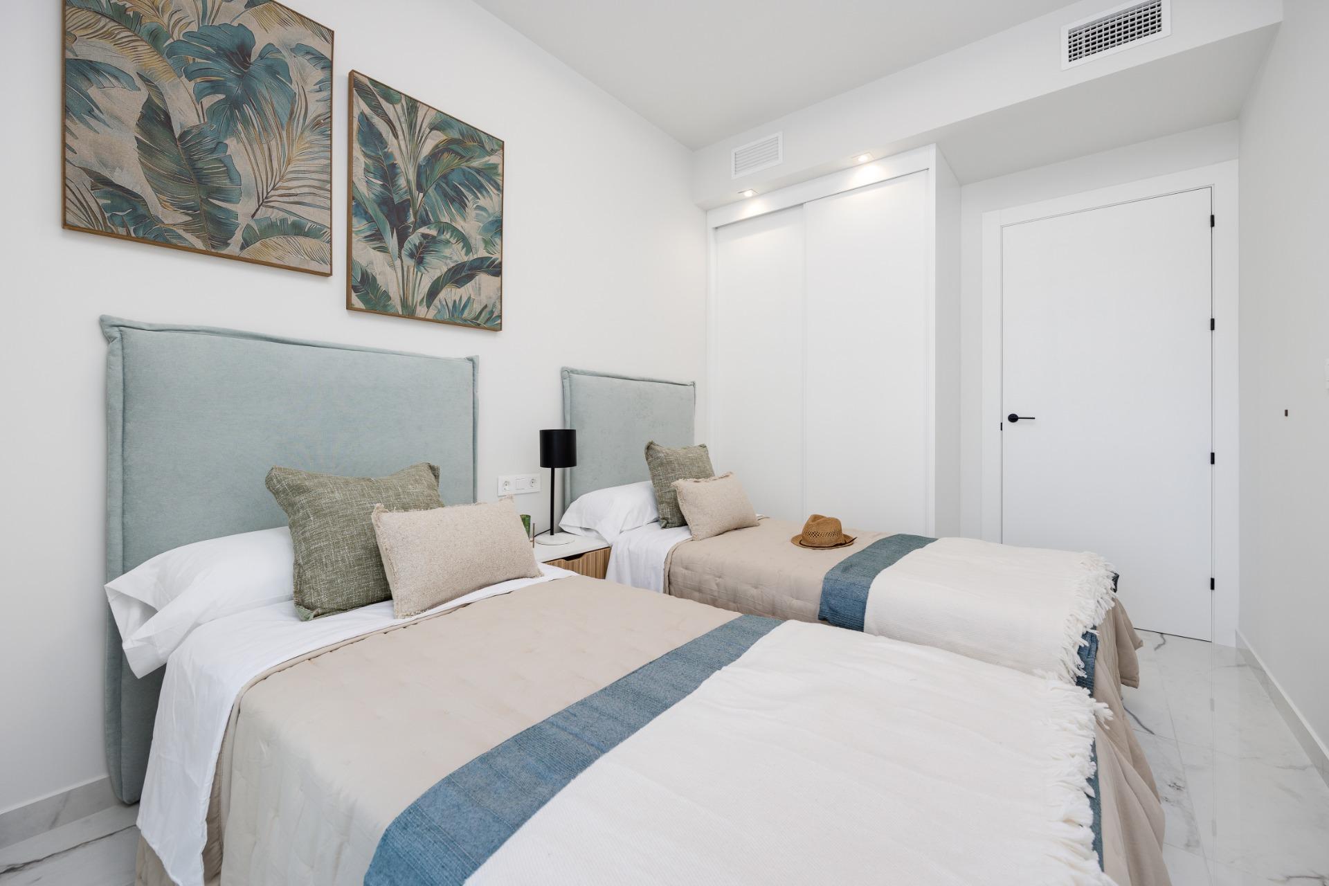 Two twin beds with light blue headboards, beige bedding, and teal accents in a bright, minimalist hotel room with tropical wall art.