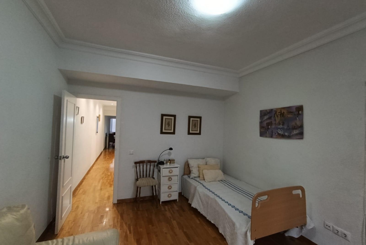 A small hospital or care-room with a single bed, nightstand, chair, and wall art, light wood floor, and an open door to a hallway.
