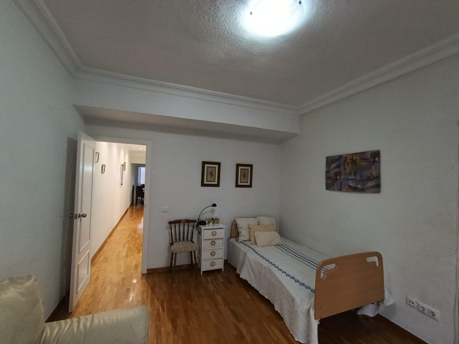 A small hospital or care-room with a single bed, nightstand, chair, and wall art, light wood floor, and an open door to a hallway.