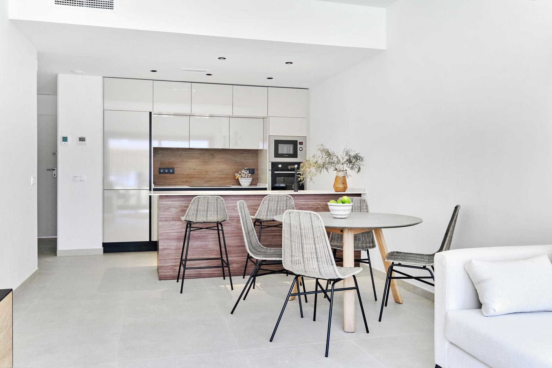 Bright open-plan kitchen with white cabinets, a wooden island, and a round dining table with wicker chairs at the island area.]','Modern kitchen: tall white cabinetry, built-in oven and microwave, wooden island, and a bowl of apples on the table.','White minimalist kitchen with stainless appliances, wooden backsplash, and four wicker stools at the island.','Dining area within kitchen: round gray table, four wicker chairs, and a vase with greenery on the counter.','Open kitchen and living area with pale tile floor, white walls, and a spacious, clean aesthetic.